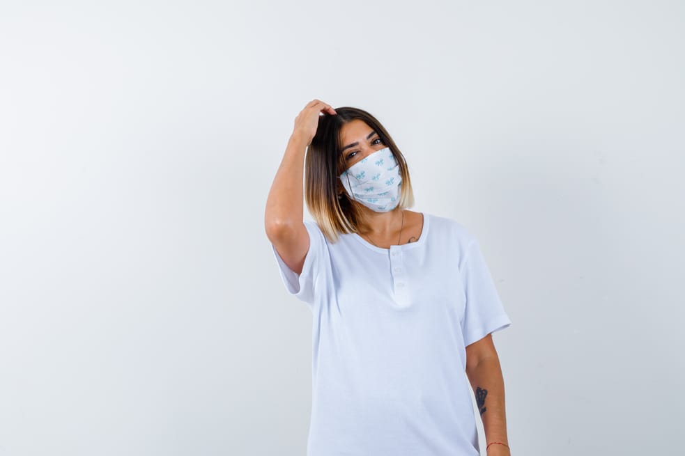 young girl scratching head in white t-shirt and mask and looking pensive. front view.
